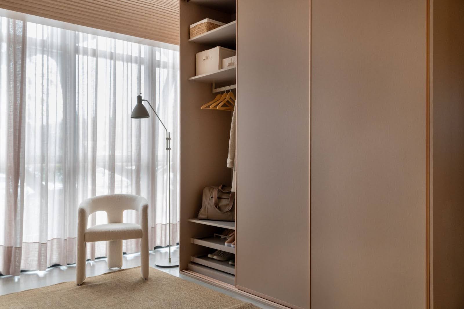 Quarto moderno com grande armário embutido de portas deslizantes, interior organizado com prateleiras e cabides, poltrona branca próxima à janela com cortinas translúcidas e luminária de chão.