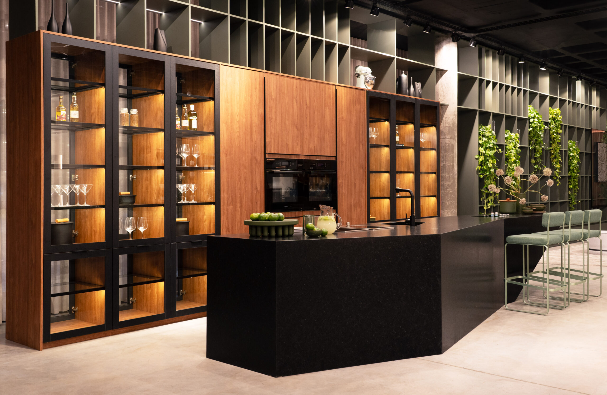 Cozinha contemporânea com ilha preta, armários altos de madeira com portas de vidro iluminadas, bancada integrada com pia e três banquetas verdes, estante aberta ao fundo com plantas e decoração minimalista.
