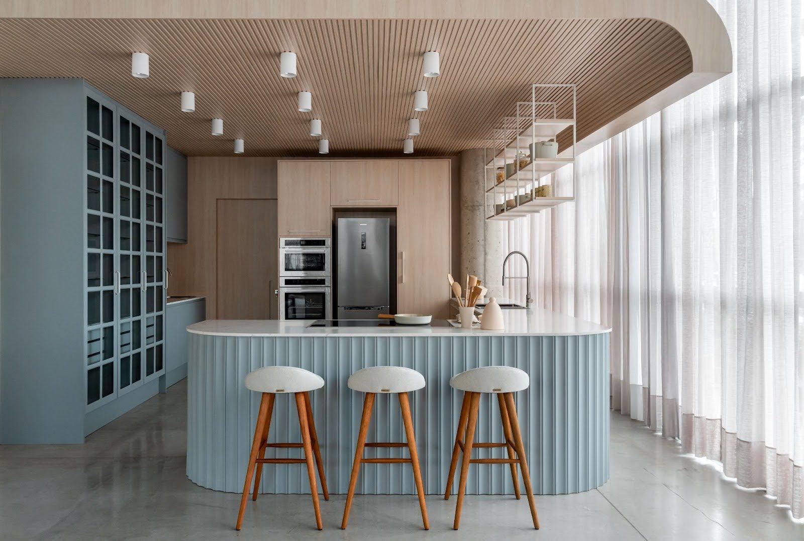 Sala de estar moderna com sofá branco, puff redondo, mesas laterais minimalistas e grande estante modular azul na parede com plantas pendentes, parede de madeira ao fundo e iluminação indireta. Sala de estar moderna com sofá branco, puff redondo, mesas laterais minimalistas e grande estante modular azul na parede com plantas pendentes, parede de madeira ao fundo e iluminação indireta.