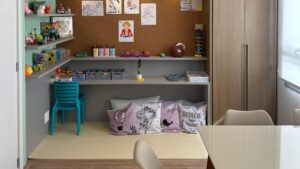 Espaço infantil com parede de cortiça decorada com desenhos, prateleiras com brinquedos e materiais de arte, cadeiras plásticas azuis, almofadas com estampas infantis e frases como “Aprecie as pequenas coisas” e “Voe alto”.