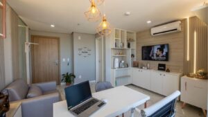 Sala com decoração moderna e aconchegante, contendo mesa branca com notebook, poltrona cinza, estante com objetos decorativos e cafeteira, TV na parede exibindo “Top Songs”, ar-condicionado e luminárias pendentes com lâmpadas expostas.