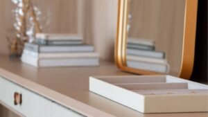 Detalhe de uma mesa de escritório ou penteadeira com livros empilhados, uma caixa de jóias com anéis dourados e um espelho de moldura dourada. O ambiente é elegante e minimalista, com uma paleta de cores suaves e um toque moderno na decoração.