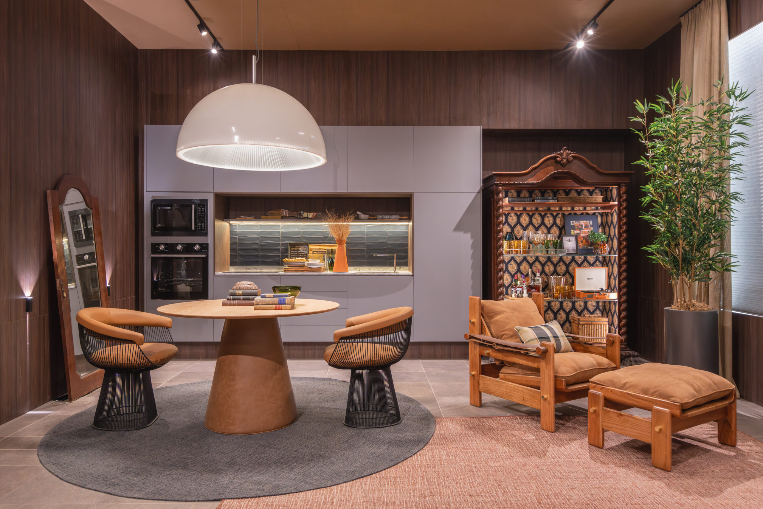 Cozinha integrada em estilo contemporâneo, com armários cinza, bancada iluminada, mesa redonda de madeira com duas cadeiras e poltronas de madeira estofadas, parede com prateleira decorativa, tapete redondo e piso de cerâmica.