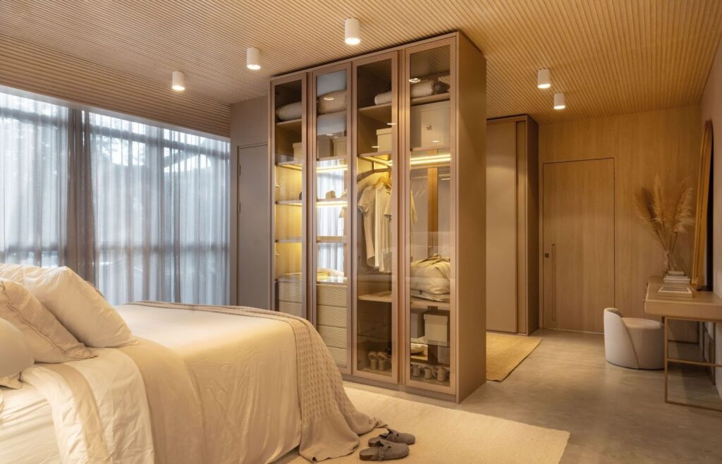 Suíte contemporânea com cama de casal, roupa de cama clara, armário com portas de vidro iluminadas internamente, espaço de circulação amplo e bancada de estudo ou penteadeira ao lado, iluminação quente e elegante.