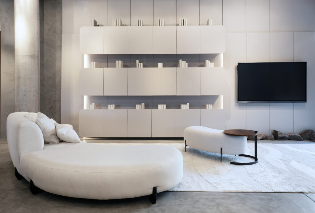 Sala de estar moderna com sofá branco curvo, poltrona branca, mesa lateral com tampo de madeira, tapete claro, parede com estante embutida branca iluminada e TV de tela plana, ambiente clean e minimalista.