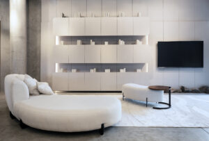 Sala de estar moderna com sofá branco curvo, poltrona branca, mesa lateral com tampo de madeira, tapete claro, parede com estante embutida branca iluminada e TV de tela plana, ambiente clean e minimalista.