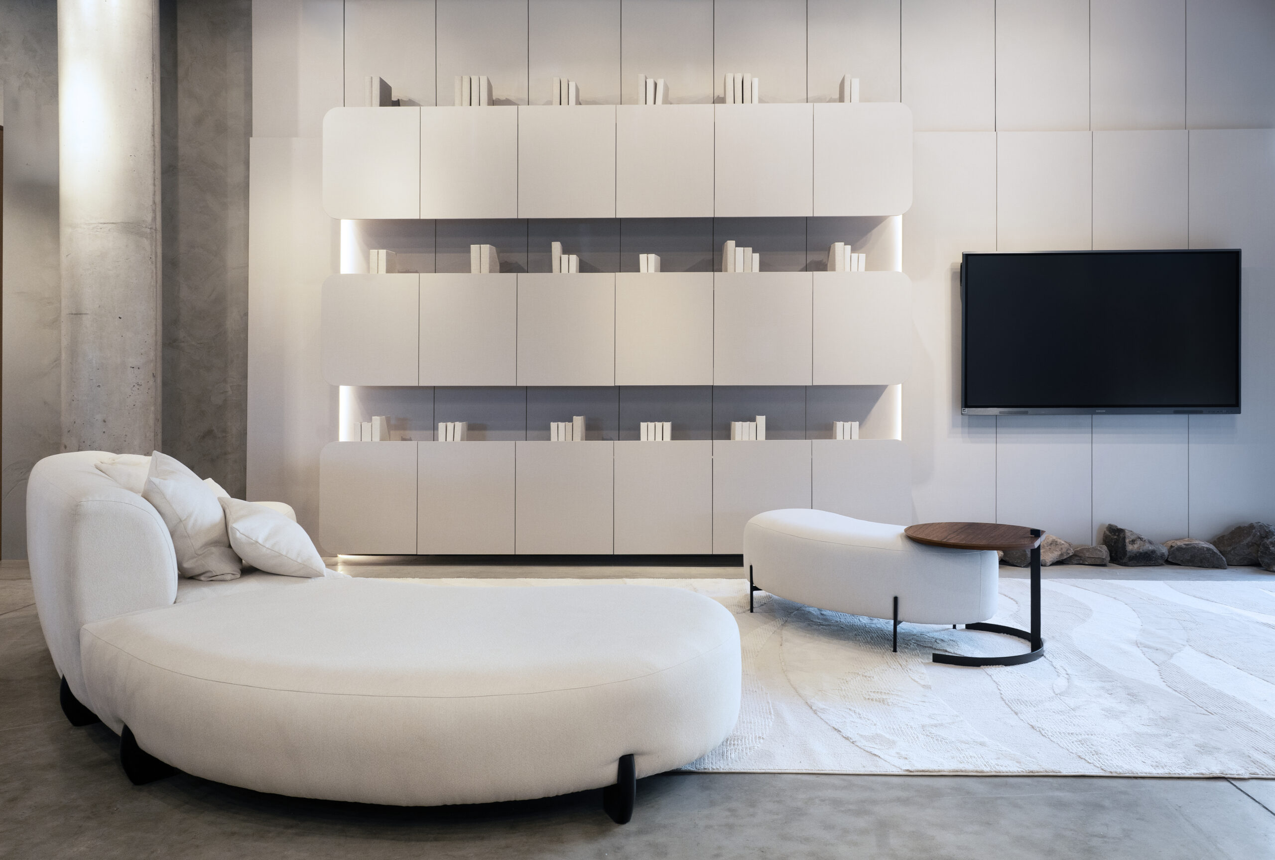 Sala de estar moderna com sofá branco curvo, poltrona branca, mesa lateral com tampo de madeira, tapete claro, parede com estante embutida branca iluminada e TV de tela plana, ambiente clean e minimalista.