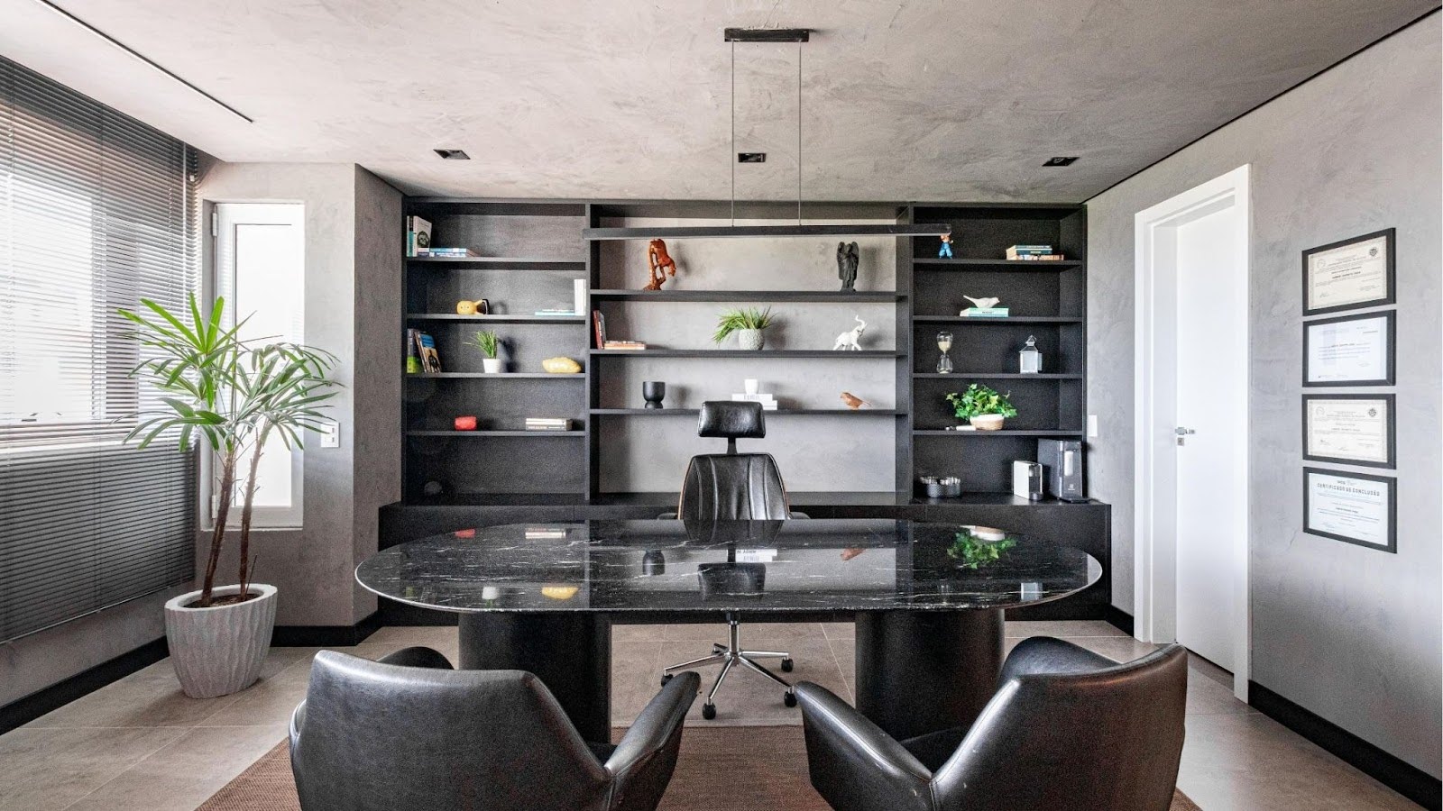 Escritório sofisticado com mesa de tampo preto brilhante, duas cadeiras de frente e uma cadeira de encosto alto ao fundo, estante preta com objetos decorativos e livros, diplomas na parede e planta em vaso próximo à janela com persiana.