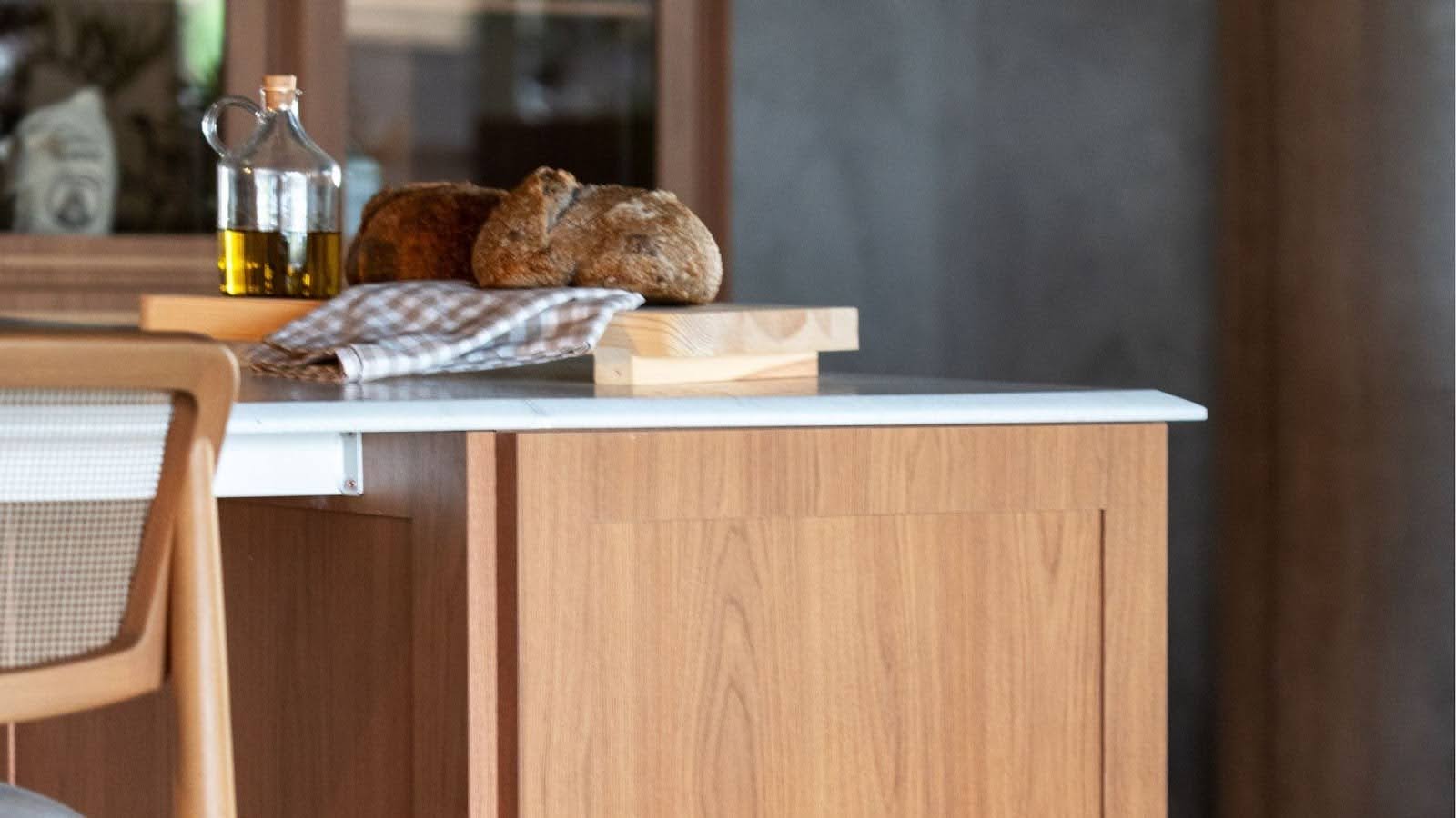 Detalhe de uma bancada de cozinha com acabamento em madeira clara. Em cima da bancada, há uma garrafa de azeite de oliva e dois pães artesanais sobre uma tábua de madeira. O ambiente é acolhedor e simples, com uma decoração clean.