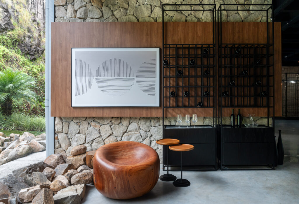 Espaço gourmet com adega vertical em metal preto e parede de pedra natural. Destaque para poltrona de madeira esculpida orgânica, painel de madeira com arte abstrata e mesas de apoio circulares.