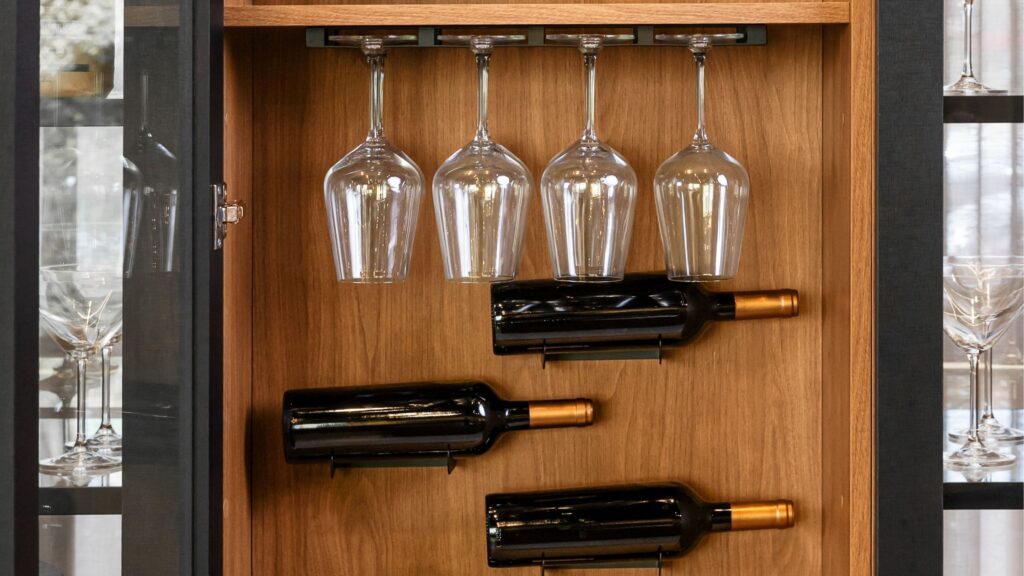 Detalhe interno de cristaleira em madeira com suporte suspenso para taças de vinho e nichos horizontais para garrafas.