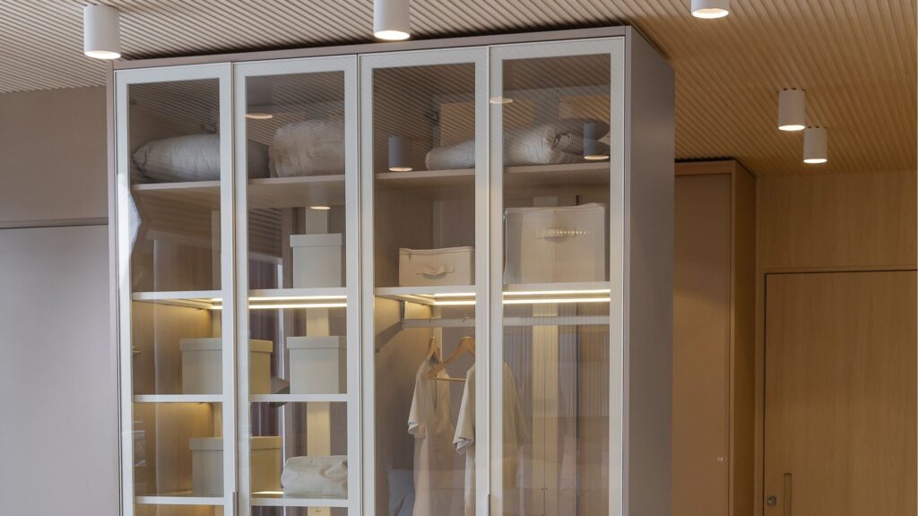 Closet planejado com portas de vidro transparente e iluminação LED interna embutida. O móvel apresenta divisórias organizadas, caixas organizadoras e teto ripado com iluminação spot.