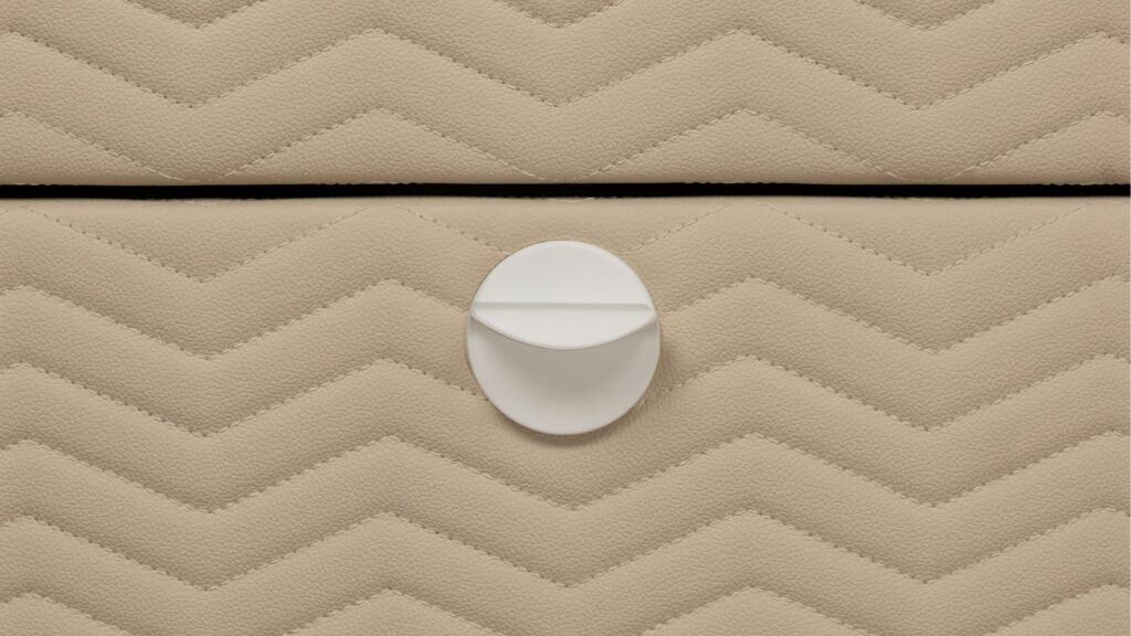 Detalhe frontal de gaveta de closet revestida em couro bege com costura matelassê em padrão zigue-zague e puxador circular branco minimalista.