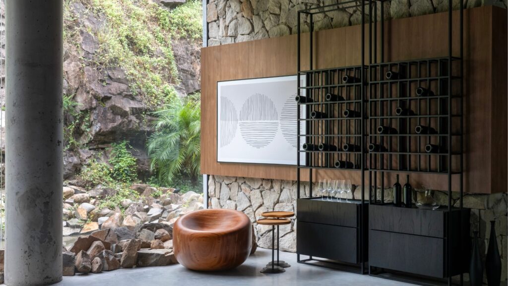 Adega de vinhos industrial preta com garrafas em parede de pedra rústica ao lado de poltrona de madeira orgânica e vista para jardim externo.