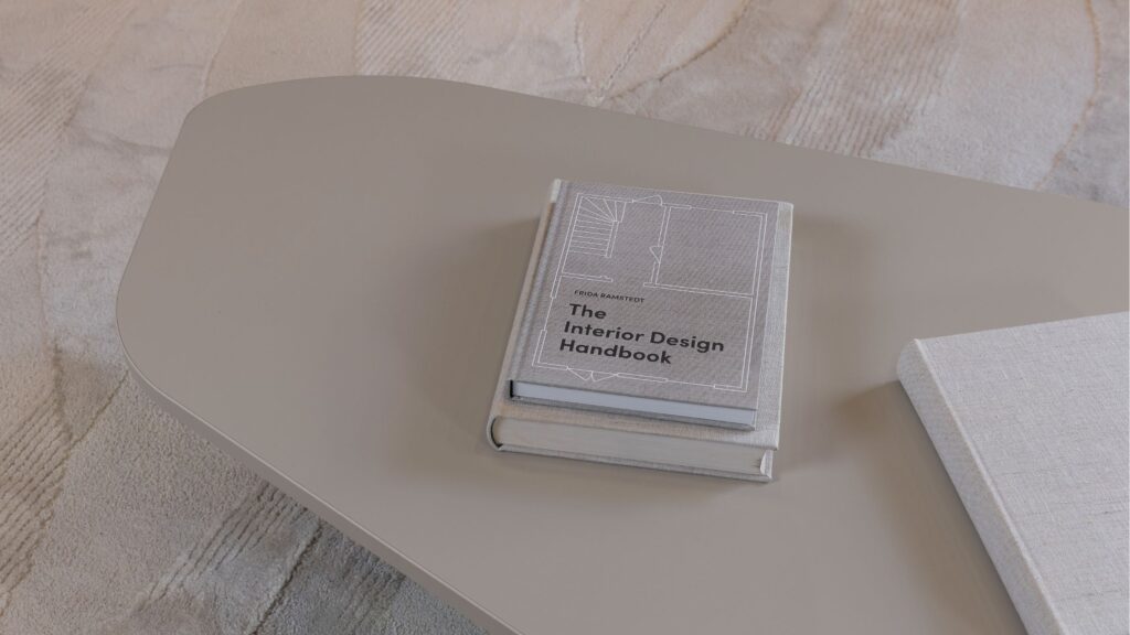Livro "The Interior Design Handbook" sobre mesa de centro orgânica bege, com tapete de textura clara em ambiente de decoração clean.